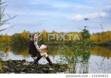 Woman painting outdoors by autumn lake on rocky shore. Woman painting outdoors by autumn lake on rocky shore. 134304633