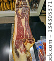 A cut of Spanish Iberico ham is placed on a stand in a supermarket. It shows marbling and color, drawing customers' attention. People browse and shop nearby. 134305371