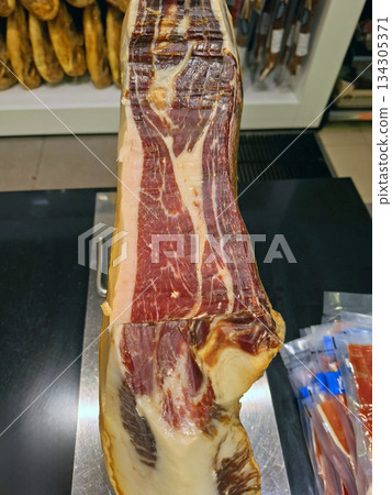 A cut of Spanish Iberico ham is placed on a stand in a supermarket. It shows marbling and color, drawing customers' attention. People browse and shop nearby. A cut of Spanish Iberico ham is placed on a stand in a supermarket. It shows marbling and color, drawing customers' attention. People browse and shop nearby. 134305371