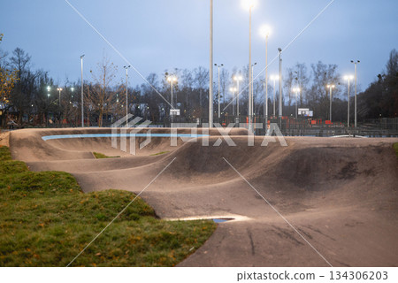 Empty outdoor asphalt pump track and dirt bike course with smooth rollers and berms illuminated by evening stadium lights. Empty outdoor asphalt pump track and dirt bike course with smooth rollers and berms illuminated by evening stadium lights. 134306203