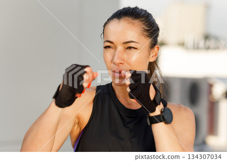 Young woman in fighting stance, ready for shadow boxing. Portrait of a young slim female wearing fitness gloves standing outdoors. Young woman in fighting stance, ready for shadow boxing. Portrait of a young slim female wearing fitness gloves standing outdoors. 134307034