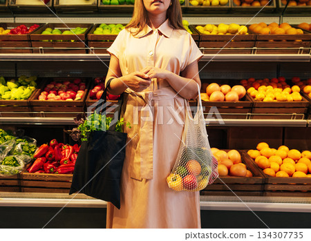 Cropped shot of a young woman with bags full of vegetables, standing in front of shelves with fruits and vegetables Cropped shot of a young woman with bags full of vegetables, standing in front of shelves with fruits and vegetables 134307735
