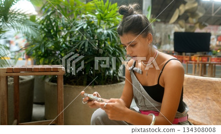 A young woman is engaged with her smartphone in a lively indoor space with vibrant decor A young woman is engaged with her smartphone in a lively indoor space with vibrant decor 134308008