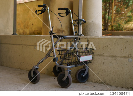 An empty walker for the elderly with a basket parked against a concrete wall outdoors. Helper for elderly and sick people 134308474
