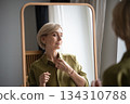 Pretty blonde middle aged woman getting ready for date, checking outlook in front of mirror at home. 134310788