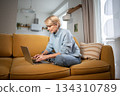 Concentrated middle aged businesswoman working comfortably with laptop on cozy couch in living room. 134310789