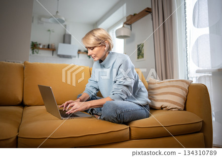 Concentrated middle aged businesswoman working comfortably with laptop on cozy couch in living room. Concentrated middle aged businesswoman working comfortably with laptop on cozy couch in living room. 134310789