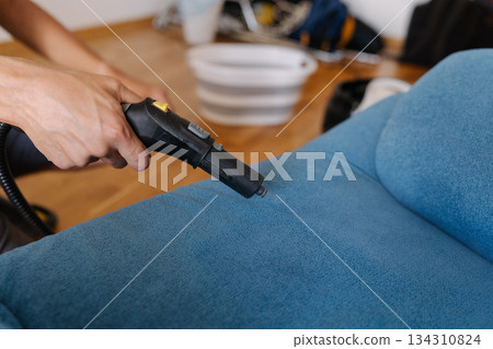 Housekeeper using steam cleaner to remove dirt and stains from blue sofa in living room, ensuring hygiene and freshness, close-up. Concept of maintenance modern home appliance. 134310824