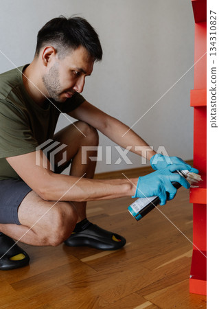Vertical portrait of young man wearing blue gloves applying scotch remover spray and scrubbing red shelf with small brush, performing thorough housekeeping at home. Concept of hygiene and cleanliness. 134310827