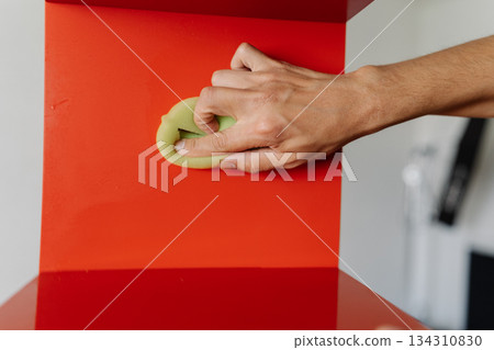 Close-up of male hand using cleaning rag, wiping dusty dirty red shelf with shiny surface, showcasing commitment to cleanliness and hygiene. Concept of domestic work, housekeeping, household. 134310830