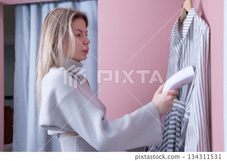 Processing woman using handheld steamer on striped shirt in home interior with neutral expression and visible steam rising from device Processing woman using handheld steamer on striped shirt in home interior with neutral expression and visible steam rising from device 134311531