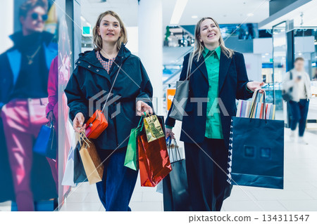 Two happy women leaving store after shopping with colorful shopping bags in their hands Two happy women leaving store after shopping with colorful shopping bags in their hands 134311547