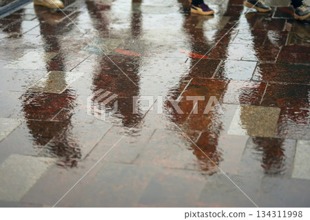 Blurry silhouettes of people reflected on wet pavement during rainy weather, footsteps visible in puddles. Concept of seasons, rainy weather, urban street reflections Blurry silhouettes of people reflected on wet pavement during rainy weather, footsteps visible in puddles. Concept of seasons, rainy weather, urban street reflections 134311998