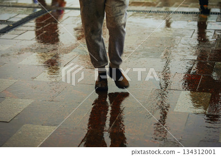 Single man walking in rain, reflected in shiny wet sidewalk with puddle. Concept of solitude, daily routine, rainy city. Abstract blurred background Single man walking in rain, reflected in shiny wet sidewalk with puddle. Concept of solitude, daily routine, rainy city. Abstract blurred background 134312001