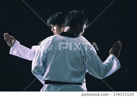 Two male martial artists practicing synchronized Taekwondo forms in traditional white uniforms on a dark background, showcasing discipline, balance, and focus in martial arts training Two male martial artists practicing synchronized Taekwondo forms in traditional white uniforms on a dark background, showcasing discipline, balance, and focus in martial arts training 134315120