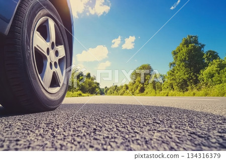 Low Angle Close-Up of Sunlit Car Tire on Asphalt Road Surrounded by Lush Greenery Low Angle Close-Up of Sunlit Car Tire on Asphalt Road Surrounded by Lush Greenery 134316379