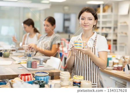 Girl stands in pottery workshop and shows created work, finished decorative vase - jug. Girl stands in pottery workshop and shows created work, finished decorative vase - jug. 134317628