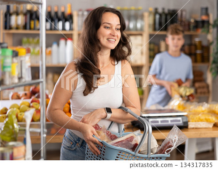Portrait of happy female shopper with cart cheese and meat choosing food and wine in grocery department of supermarket 134317835