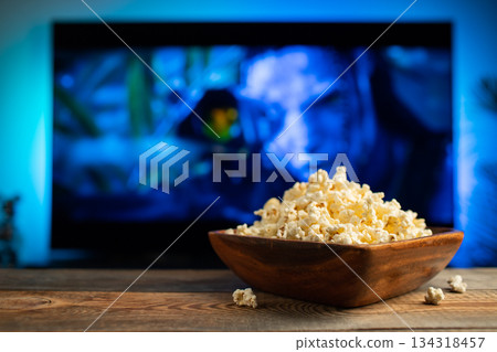 A wooden bowl of popcorn and remote control in the background the TV works. Evening cozy watching a movie or TV series at home A wooden bowl of popcorn and remote control in the background the TV works. Evening cozy watching a movie or TV series at home 134318457