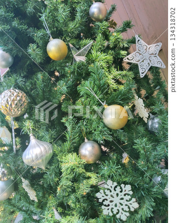 Decorated Christmas tree with ornaments and decorations. The ornaments include golden spheres, silver bulb-shaped objects, snowflake-shaped objects, and a star-shaped ornament 134318702