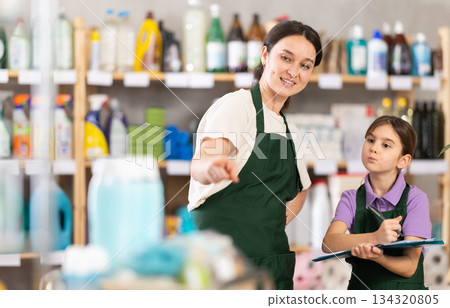 Mother teaches little daughter to keep track of goods in the store. Daughter makes notes on paper in folder Mother teaches little daughter to keep track of goods in the store. Daughter makes notes on paper in folder 134320805
