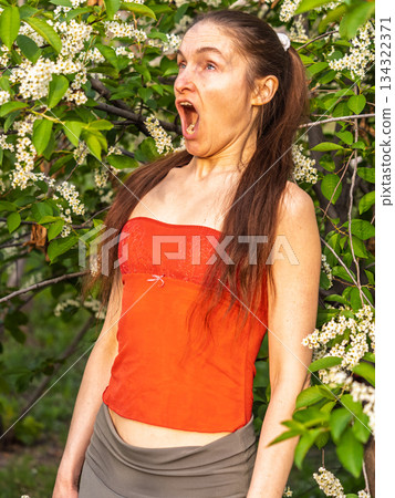 Young woman sneezes in the park against the background of a flowering tree. Allergy to pollen concept. Young woman sneezes in the park against the background of a flowering tree. Allergy to pollen concept. 134322371