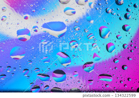 Abstract macro photography of water droplets on a vibrant neon blue and purple gradient background, creating a psychedelic liquid texture pattern. 134322599