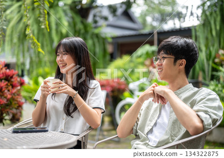 Pretty asian woman holding coffee and talking with glasses man while sitting at table in cafe garden 134322875