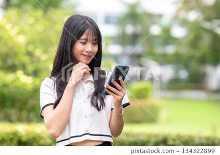 Pretty asian woman with hand under chin looking at phone while standing in green garden cafe or park Pretty asian woman with hand under chin looking at phone while standing in green garden cafe or park 134322899