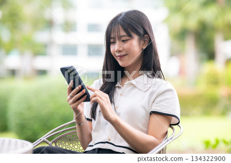 Pretty asian woman typing or looking at phone while sitting on chair at table in garden cafe or park Pretty asian woman typing or looking at phone while sitting on chair at table in garden cafe or park 134322900