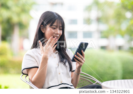 Pretty asian woman looking at phone in surprised while sitting on chair at table in a garden or park 134322901