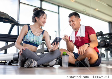Asian woman client showing phone to fitness coach trainer man sits on floor after exercising in gym. 134324081