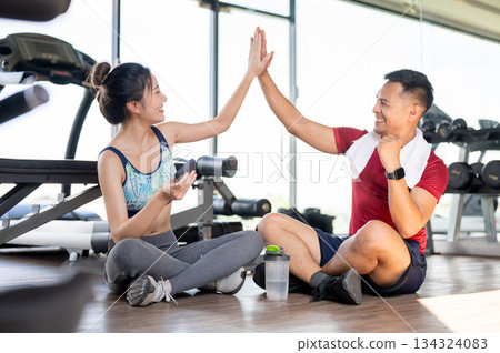 Asian woman client and fitness coach clapping hands high five sits on floor after exercising in gym 134324083