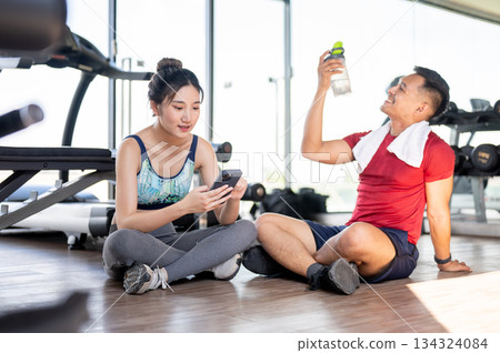 Woman client looking at phone and fitness gym trainer drinking water sits on floor after exercising. 134324084