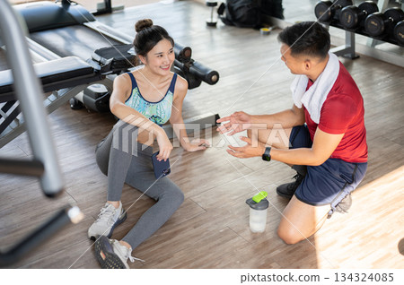 Woman client holding phone talking with fitness trainer as sitting on floor after exercising in gym. Woman client holding phone talking with fitness trainer as sitting on floor after exercising in gym. 134324085