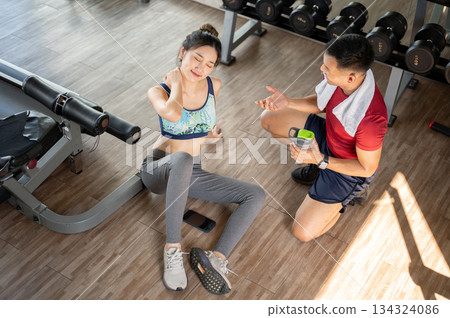 Woman client massaging her neck aside fitness trainer while sitting on floor after exercising in gym 134324086