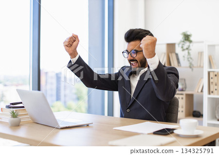 An Indian businessman celebrates a victory in his office, looking at his laptop with excitement An Indian businessman celebrates a victory in his office, looking at his laptop with excitement 134325791