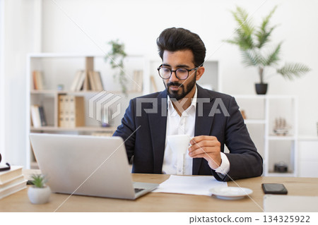 An Arabian businessman is working on his laptop while drinking coffee in his office An Arabian businessman is working on his laptop while drinking coffee in his office 134325922