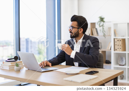 An Arab businessman enjoys a coffee break while working on his laptop in a modern office setting An Arab businessman enjoys a coffee break while working on his laptop in a modern office setting 134325924