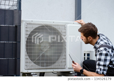 Technician servicing outdoor air source heat pump unit near house wall 134326179