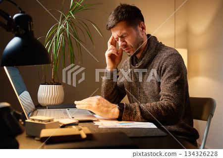 Fatigued man with headache sitting at desk while working at home Fatigued man with headache sitting at desk while working at home 134326228