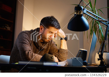 Fatigued man sitting at home desk looking at laptop Fatigued man sitting at home desk looking at laptop 134326229