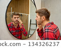 Man cleansing face with cotton pad in front of mirror in bathroom 134326237