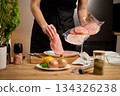 Woman placing raw chicken fillets from packaging onto a plate in home kitchen 134326238