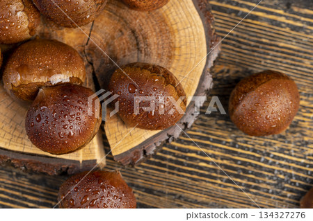 a hard edible chestnut fruit in a brown shell covered with water droplets on a wooden table, a group of wet and fresh chestnut fruits with a brown hard shell 134327276
