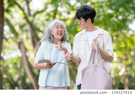 Old woman grandmother laughing after reading diary book as walking with young man grandson in park. Old woman grandmother laughing after reading diary book as walking with young man grandson in park. 134327414