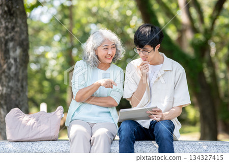 Old woman grandmother pointing and young man grandson looking at tablet as sitting on bench in park. Old woman grandmother pointing and young man grandson looking at tablet as sitting on bench in park. 134327415