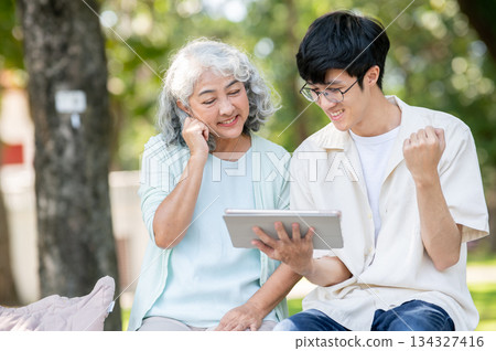 Old woman grandmother looking at tablet and young man grandson making a fist while sitting in park. Old woman grandmother looking at tablet and young man grandson making a fist while sitting in park. 134327416
