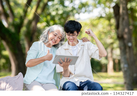 Thumb up old woman grandmother looking at tablet and young man grandson raising a fist sits in park. Thumb up old woman grandmother looking at tablet and young man grandson raising a fist sits in park. 134327417