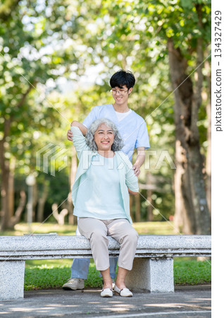 Glasses man caregiver nurse helping old woman granny patient exercise while sitting on bench in park Glasses man caregiver nurse helping old woman granny patient exercise while sitting on bench in park 134327429